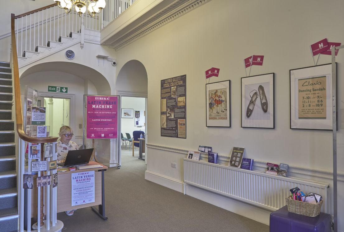 A women sat at a desk in the downstairs area of a building. To her left is the start of the staircase, to the right displays and HODs flags!