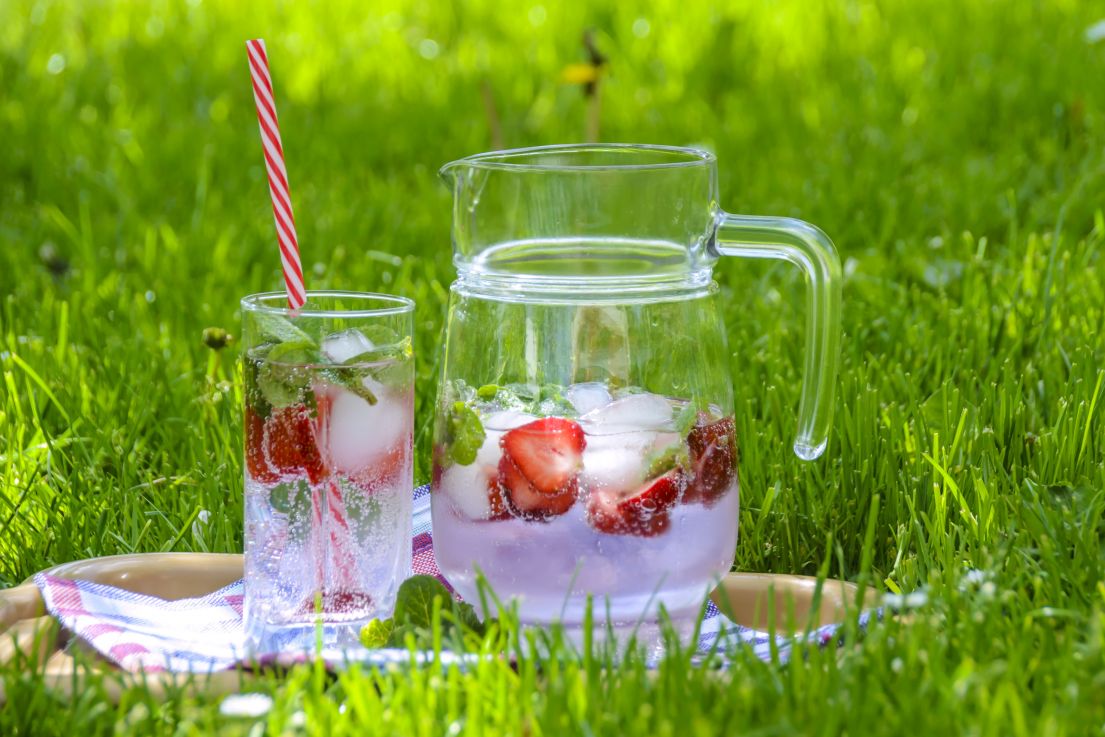 Placed on the grass is a jug of a clear fizzy drinks with floating strawberries, next to a filled glass with a long red stepped straw.