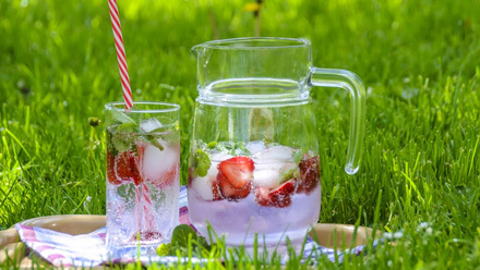 Placed on the grass is a jug of a clear fizzy drinks with floating strawberries, next to a filled glass with a long red stepped straw.