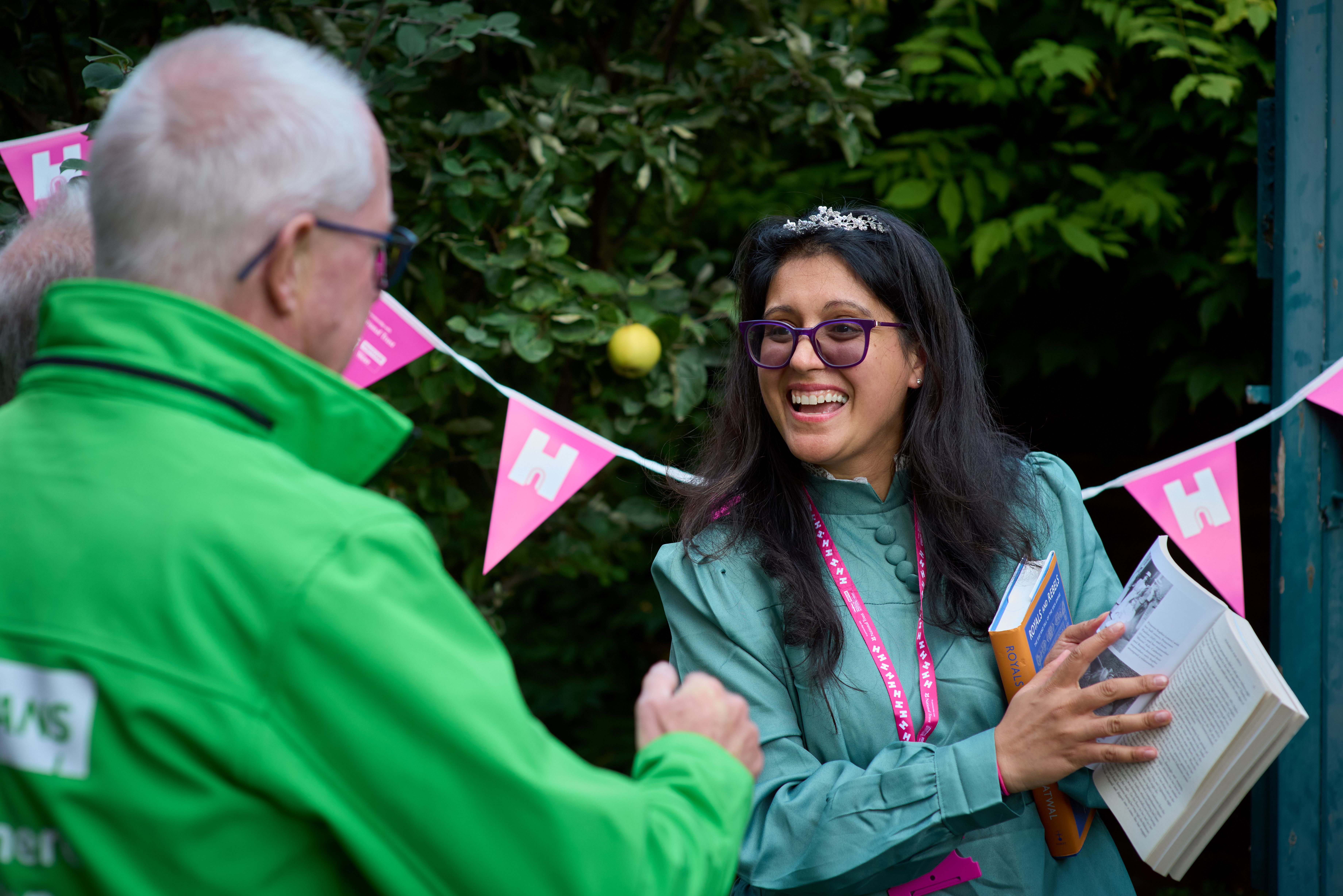 Stood in front of a hedge with bunting hung up, a young woman showing an older man a picture in a book.