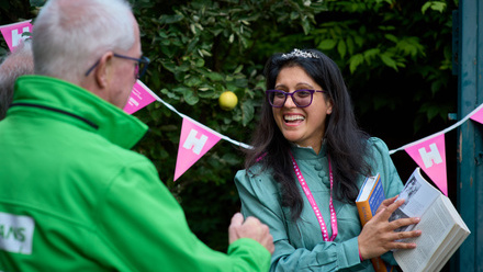 Stood in front of a hedge with bunting hung up, a young woman showing an older man a picture in a book.