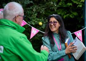 Stood in front of a hedge with bunting hung up, a young woman showing an older man a picture in a book.