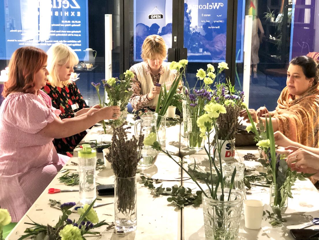 Four women sat around a long table, arranging flowers that are available in vases dotted around the table.