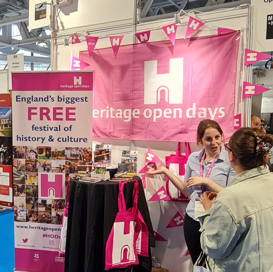 An exhibition stall for Heritage Open Days. A women with tied up hair talks to a stall visitor, behind them are a variety of HODs publicity materials 