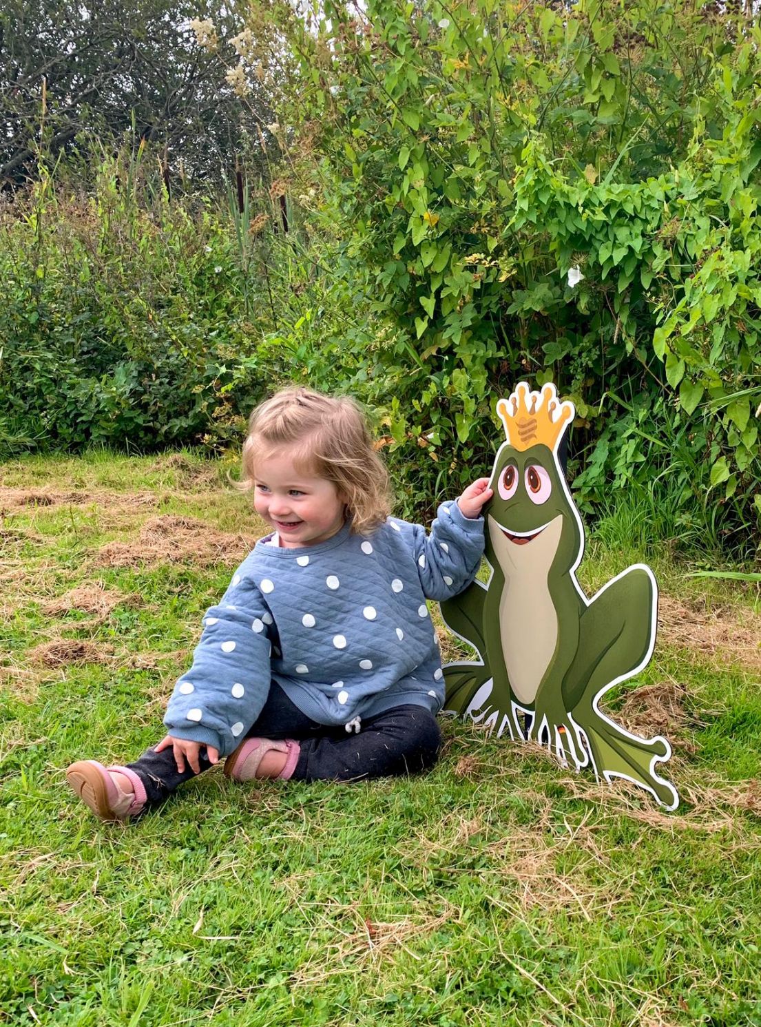 A small little girl wearing a blue and white pokadot jumper and jeans, smiling and sat next to a frog king cardboard cut out.