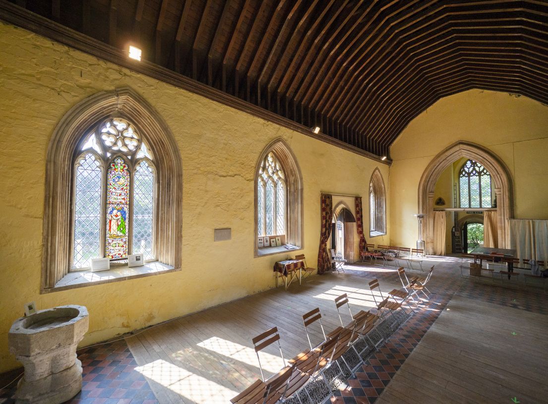 Inside of a wooden vaulted ceiling hall, with yellow stone walls and arched windows. To left is a baptism font and chairs lining the centre.