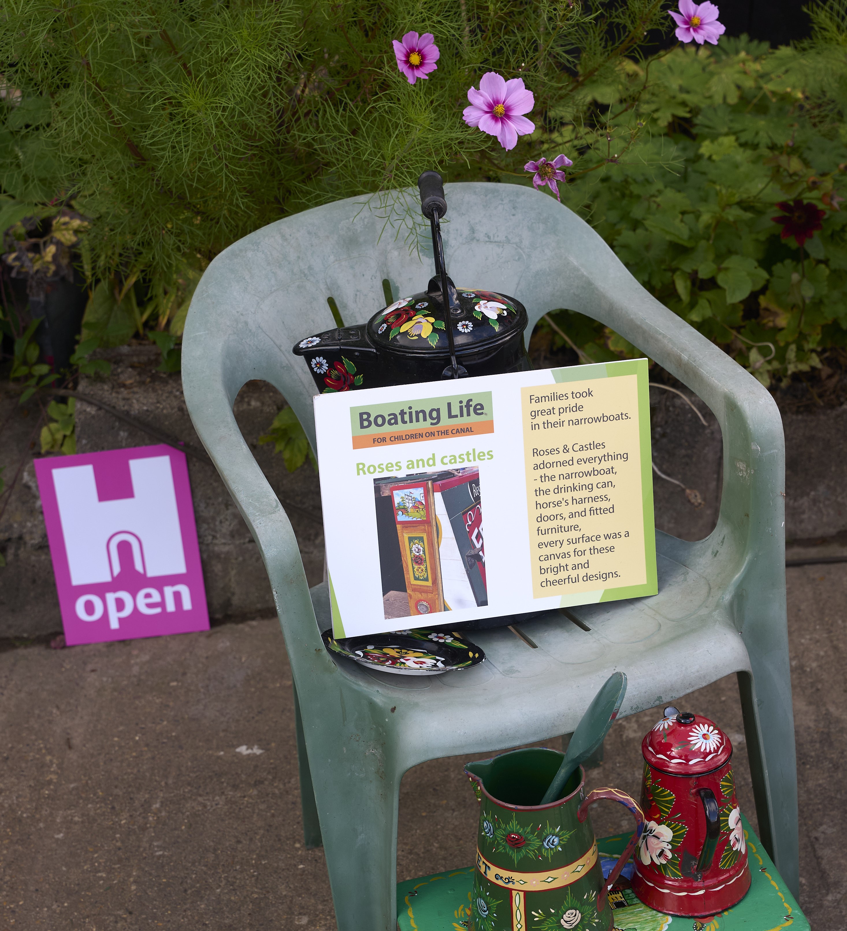 Interpretation board on a plastic garden chair with decorated canal tin jugs in front of it.