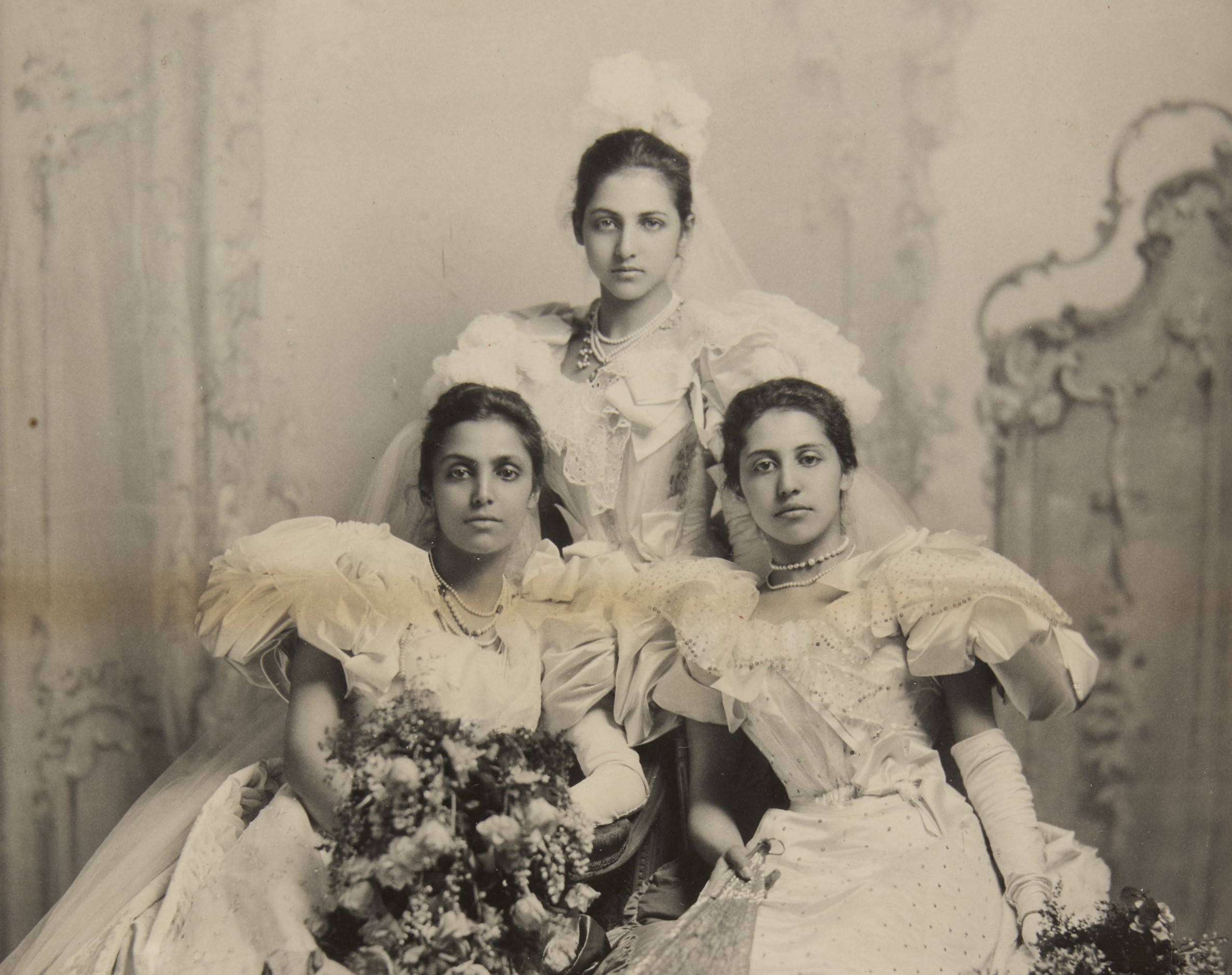 Posed group photograph of three young Asian women in grand white Victorian dresses.