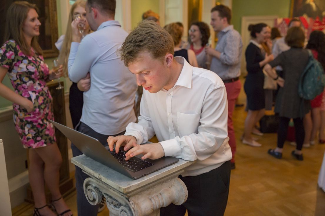 A man in a white shirt and dark trousers typing on a laptop on a plinth. Behind him, people are networking. 