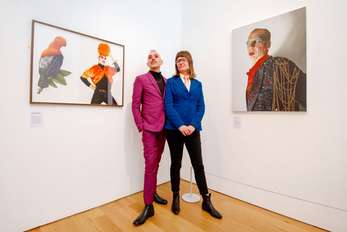 Two individuals standing in-front of artwork. On the left a man with grey hair & a dark pink suit, on the right a lady with brown hair and blue suit.