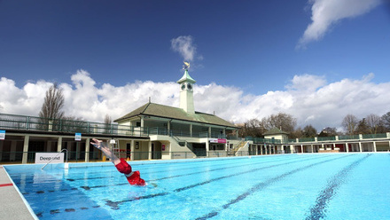 Person in red bathing suit diving into deep end of outdoor pool, surrounded by changing rooms and a clocktower building.