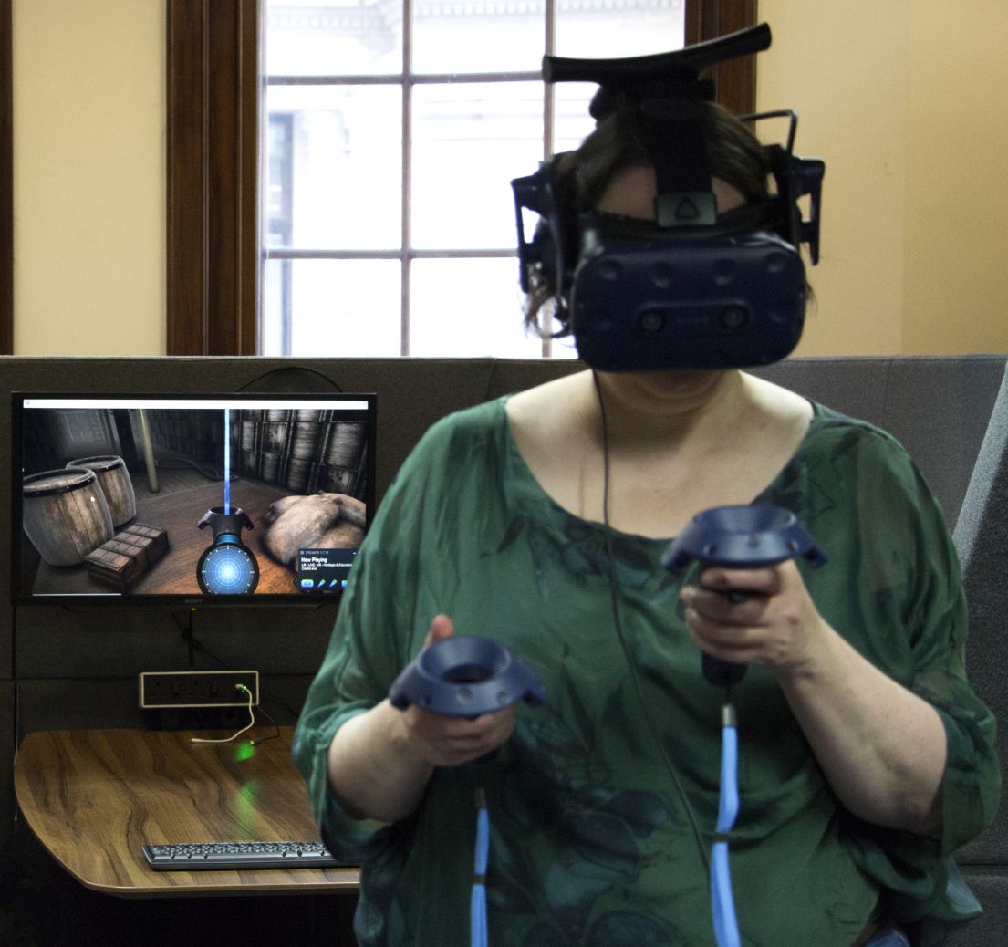 A women with a green scooped t-shit, wearing a VR head set, with interactive remotes in her hands. Behind her is a screen showing what she is seeing.