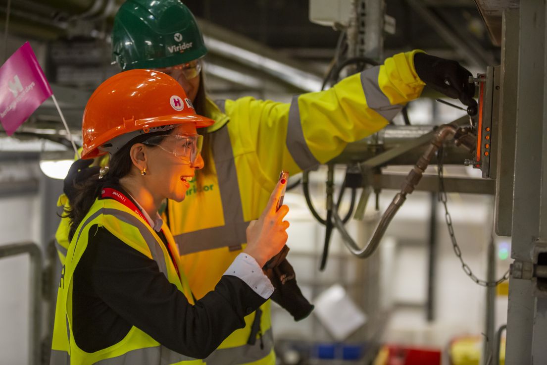 Two women wearing hard hats, one holding a HODs hand held flag and opening a viewing hatch to an incinerator. The other takes a photo on her phone.