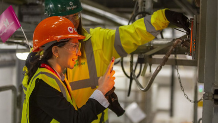 Two women wearing hard hats, one holding a HODs hand held flag and opening a viewing hatch to an incinerator. The other takes a photo on her phone.