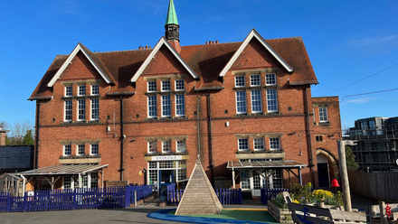 Large red brick gabled building with a narrow central spire, and a playground in front of the entrance.