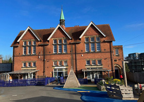 Large red brick gabled building with a narrow central spire, and a playground in front of the entrance.
