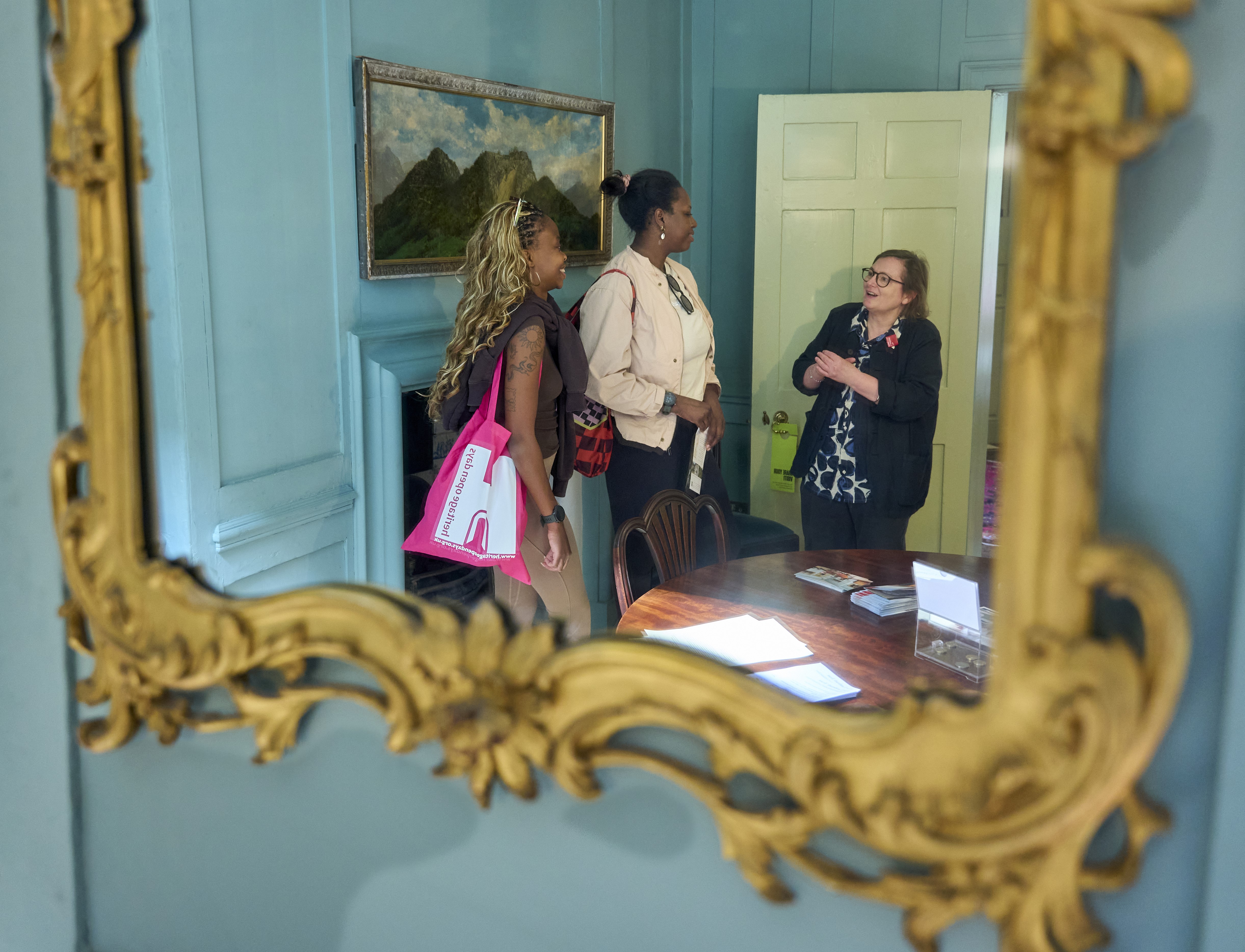 Two visitors chatting to a volunteer in a wood panelled room - reflected in a gilt mirror.