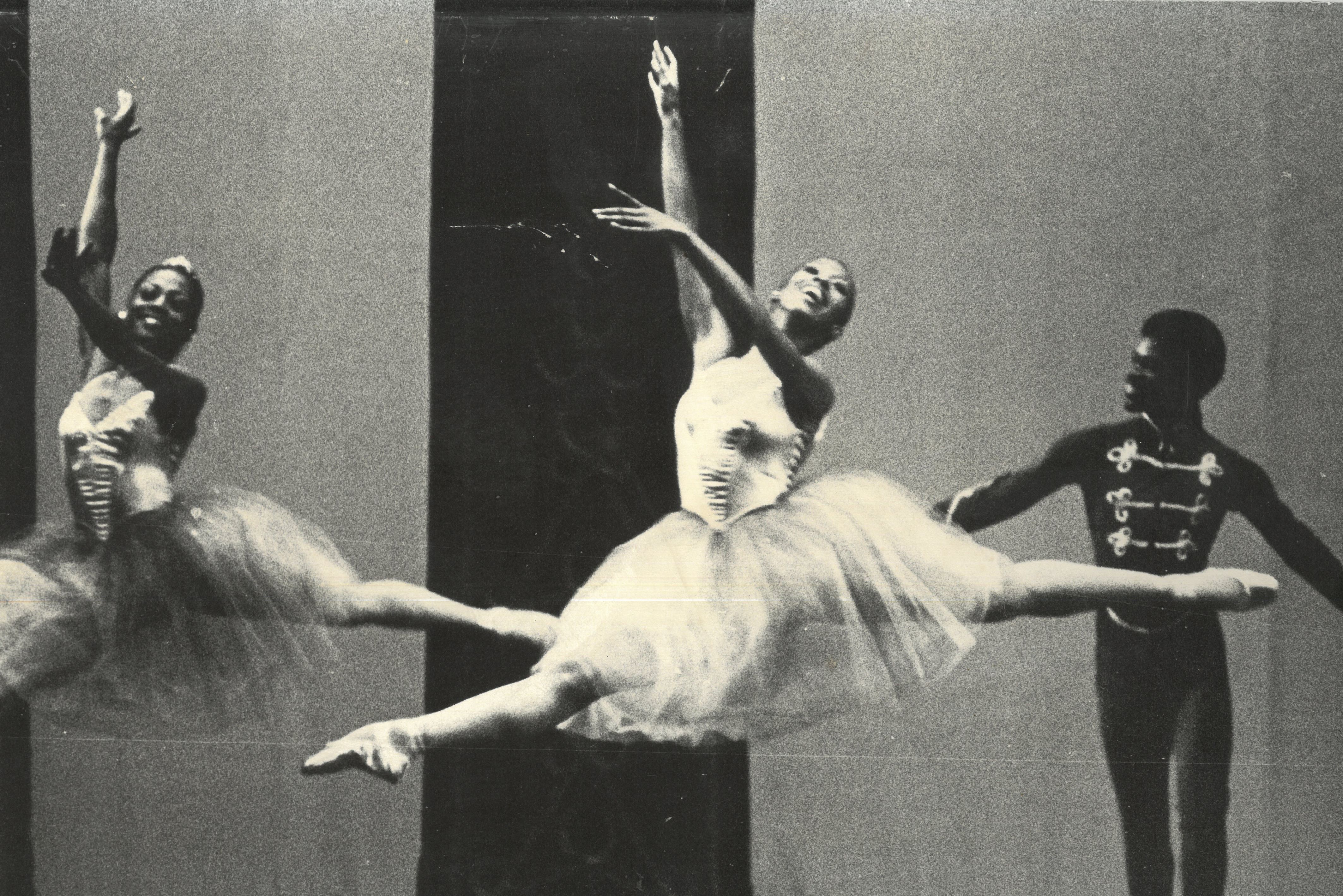 Two female Black ballet dancers leap across a stage, while a male dancer stands behind.