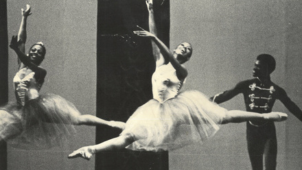 Two female Black ballet dancers leap across a stage, while a male dancer stands behind.