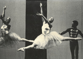 Two female Black ballet dancers leap across a stage, while a male dancer stands behind.