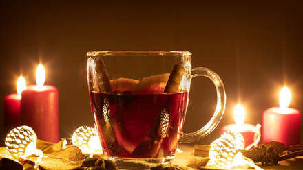 A glass mug of a red drink, with orange slices and cinnamon sticks. Around the mug are fairly lights and candles.