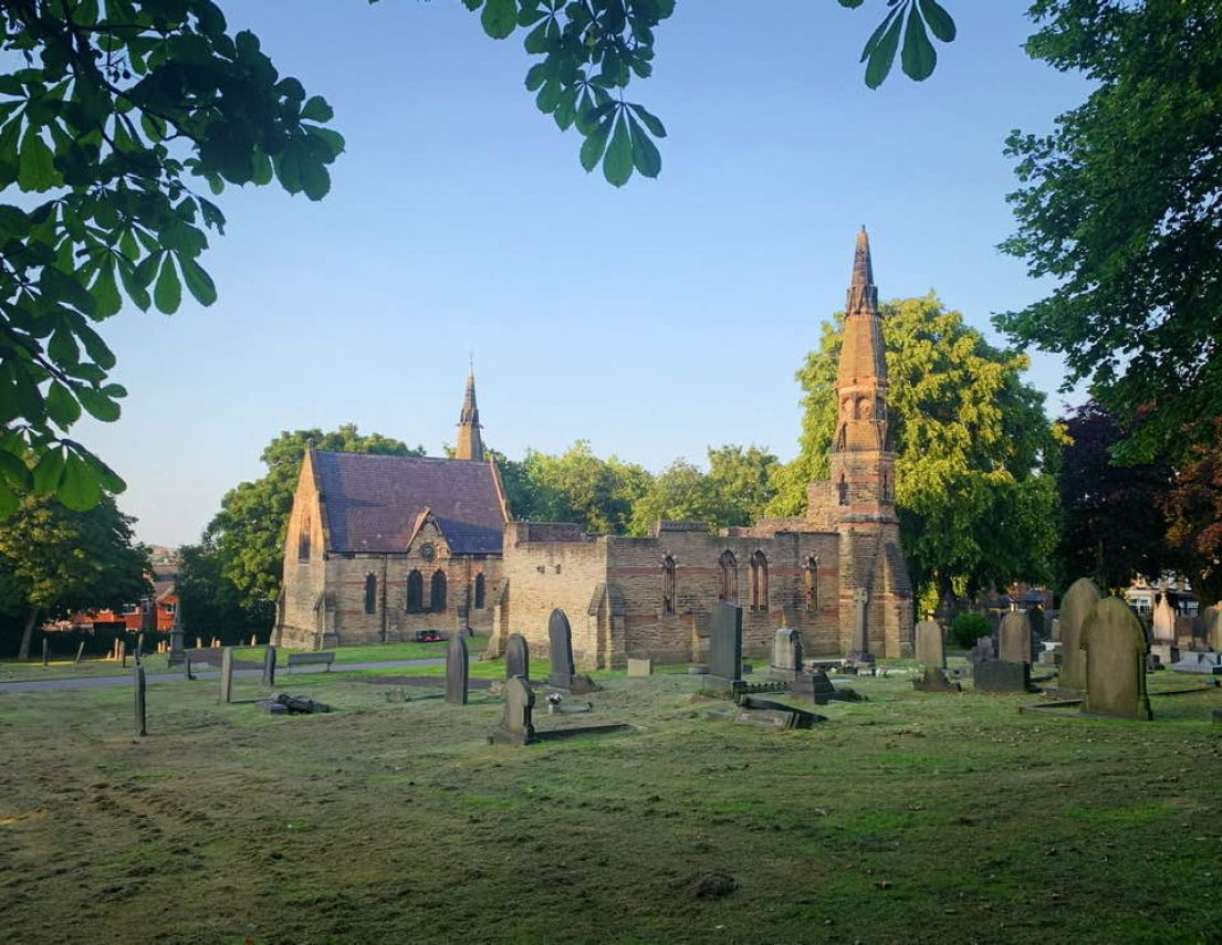 Looking on from a distance, a church set within a large field, filled with different shaped headstones. Around the perimeter are tall green trees. 