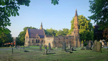 Looking on from a distance, a church set within a large field, filled with different shaped headstones. Around the perimeter are tall green trees. 