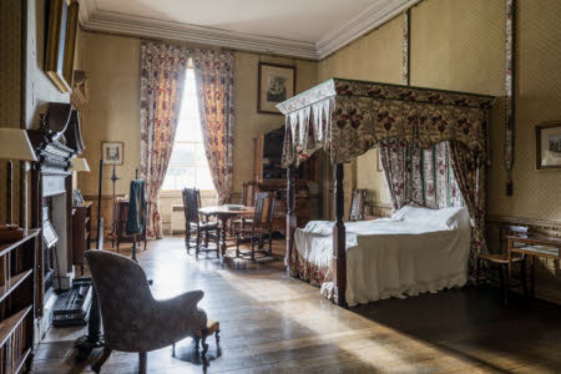 A large bedroom, with a four poster bed, a table and chairs and several photographs on the wall and a chair next to the fireplace.