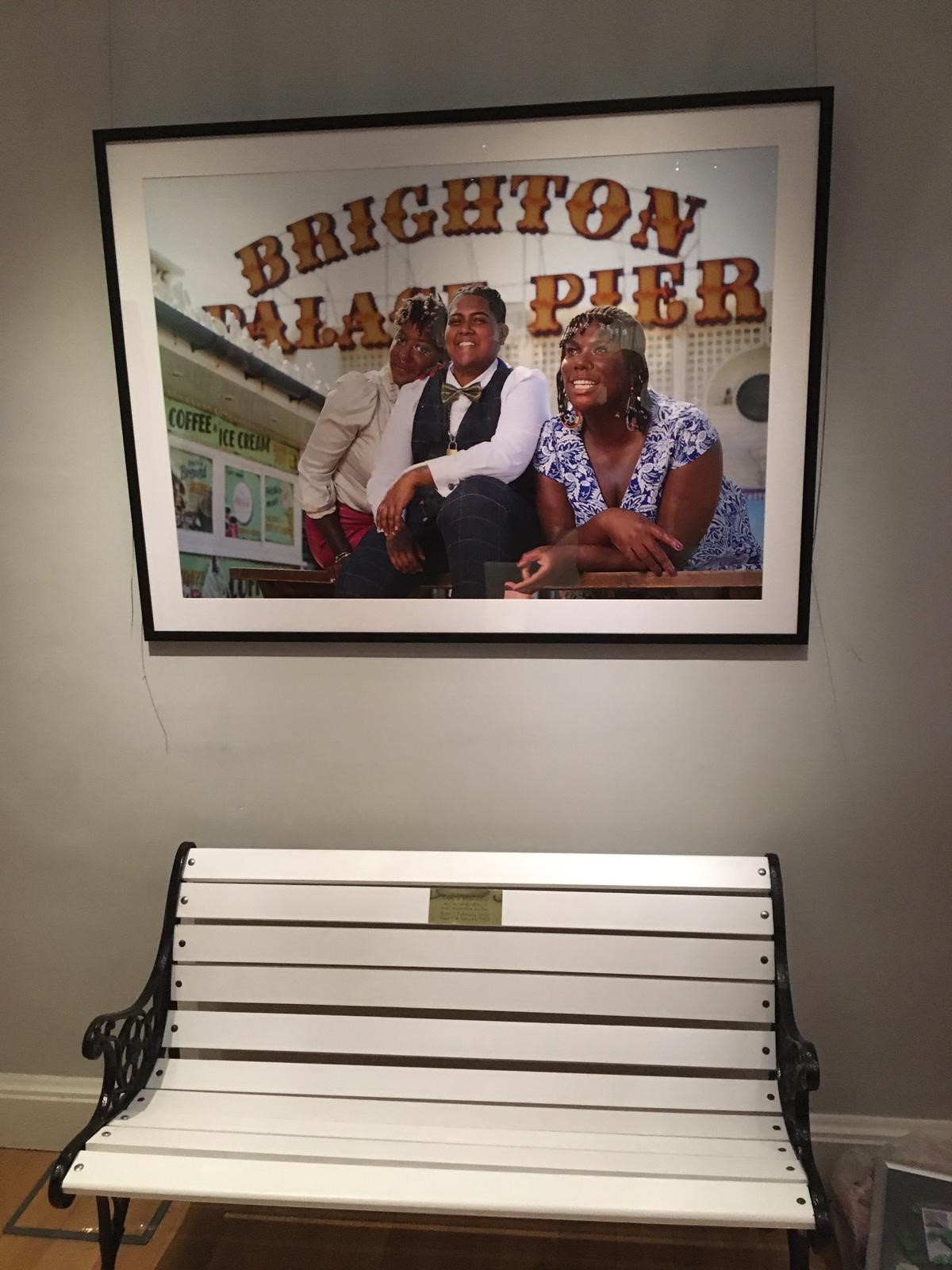 Framed photo of three people on Brighton pier hanging above a white bench with black metal arms in a room.