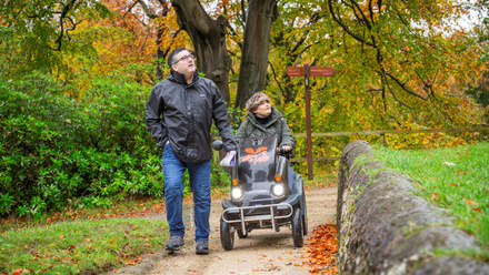 Man walking beside a woman driving a mobility vehicle along a paved path in Autumnal parkland.