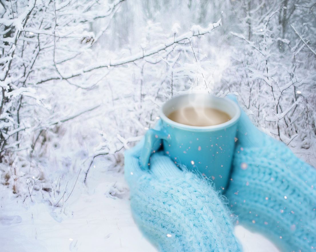 A person with light blue mittens holding a steaming mug of tea in-front of a snowy background.