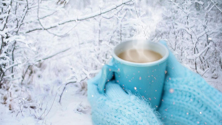 A person with light blue mittens holding a steaming mug of tea in-front of a snowy background.