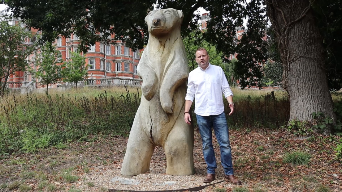 A man in a white shirt and dark jeans standing next to a wooden carving of a bear, standing on its hind paws.