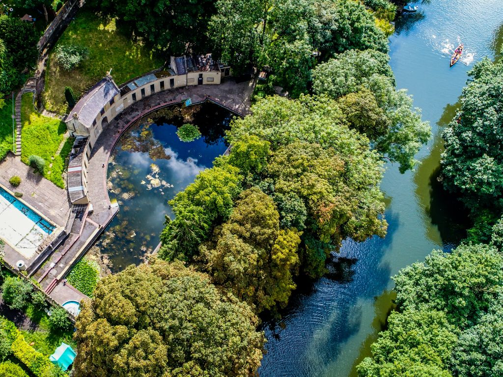 Aerial view of river bordered by trees and a semi-circular pool bordered by a curved building.