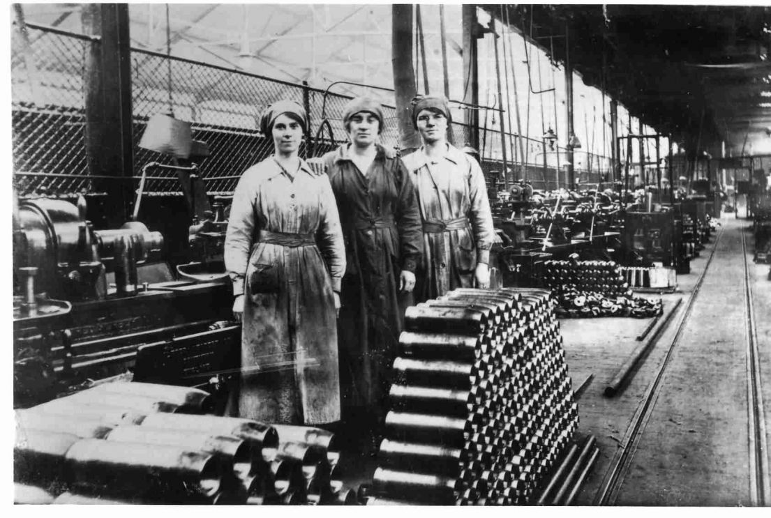 A black and white image of three women dressed in work overalls in a factory. They are surrounded my stacked ammunition. 