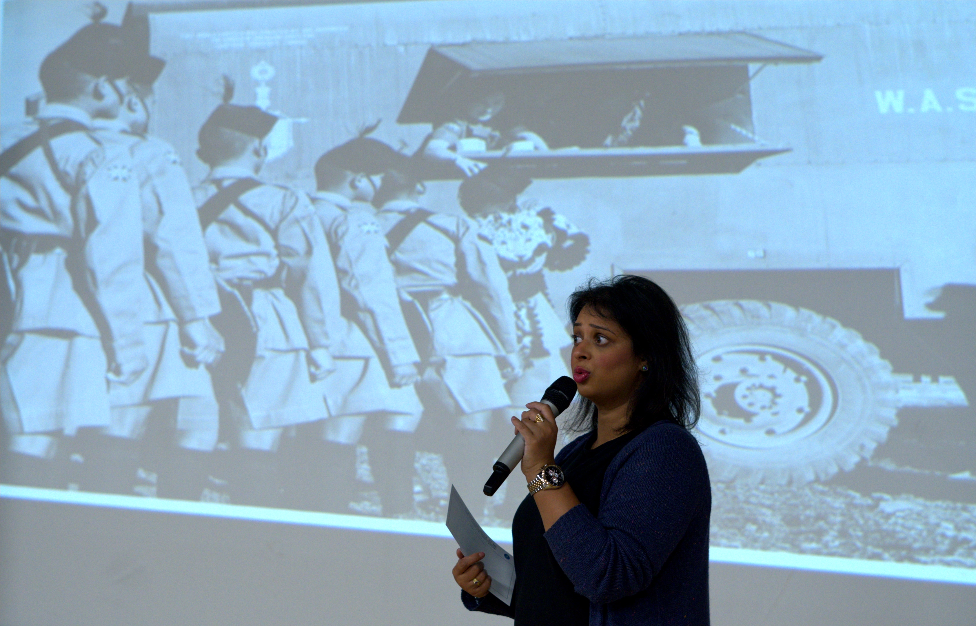 Woman speaking into a microphone in front of a large digital sceen displaying a wartime photo of soldiers queueing at a canteen truck.