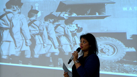 Woman speaking into a microphone in front of a large digital sceen displaying a wartime photo of soldiers queueing at a canteen truck.