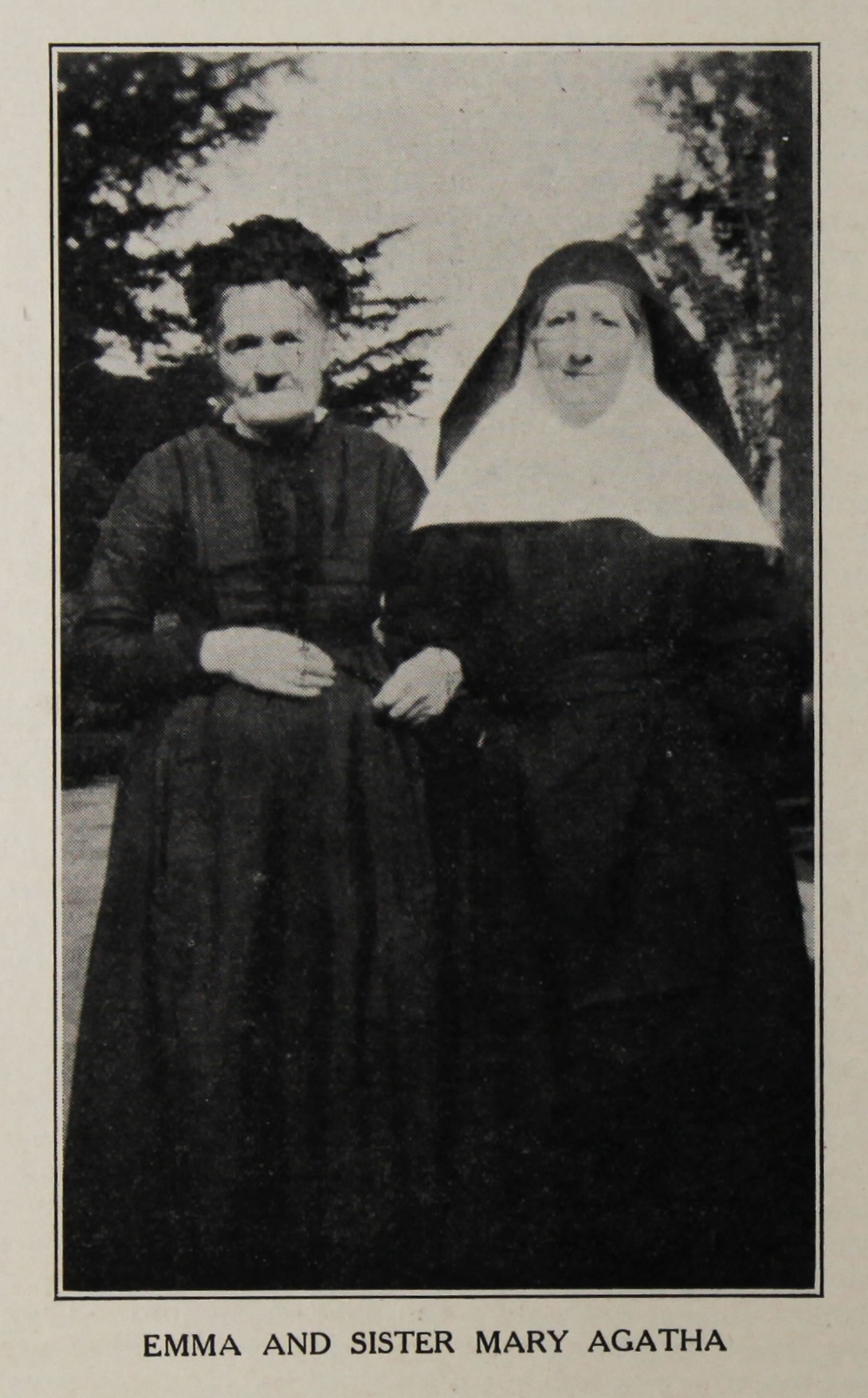 Photograph of elderly woman in a black dress stood outside arm in arm with a nun.