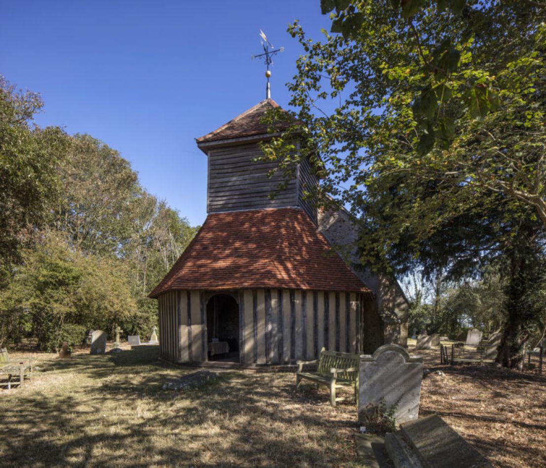 A wooden timbered church in a church yard, surrounded by gravestones and trees.