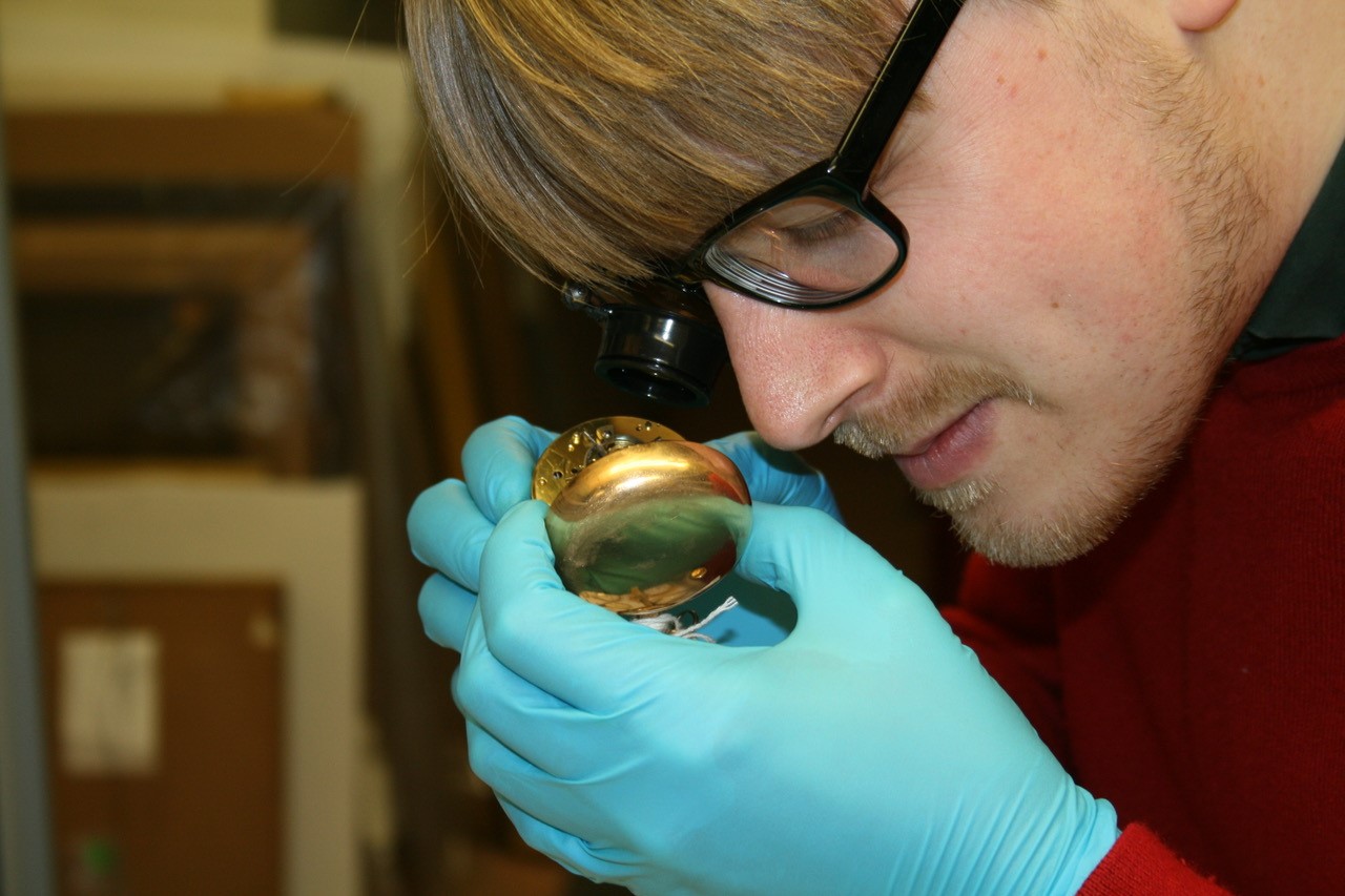 Man wearing gloves and  an eyepiece holds a pocketwatch close to see inside.