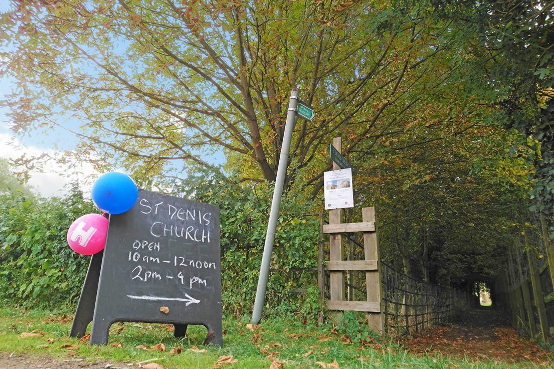 A slate sign pointing to a public foot path. On the sign written in chalk reads 'St Denis Church Open 10am-12 noon & 2pm-4pm'.