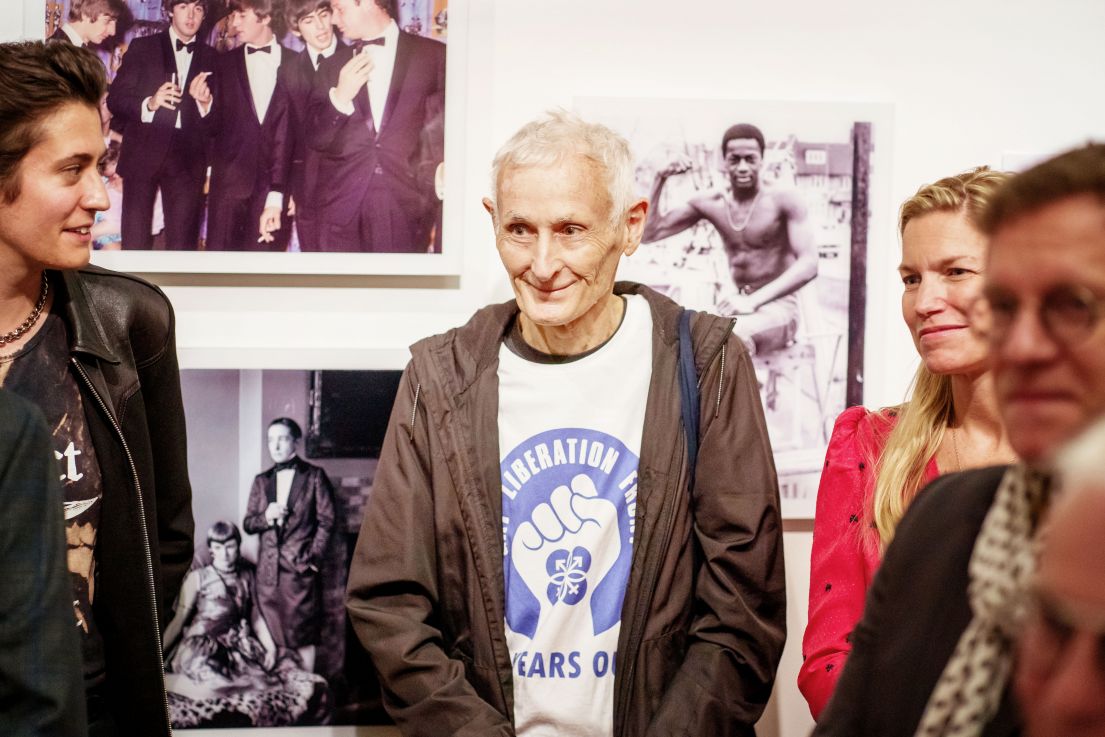 A group of people surrounding a man with white hair, brown coat, wearing a White T-shirt with Gay Liberation Front in Blue on the front.