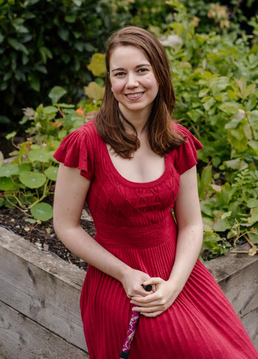 Younger woman in red dress, holding a cane,  seated on the corner of a wooden planter.