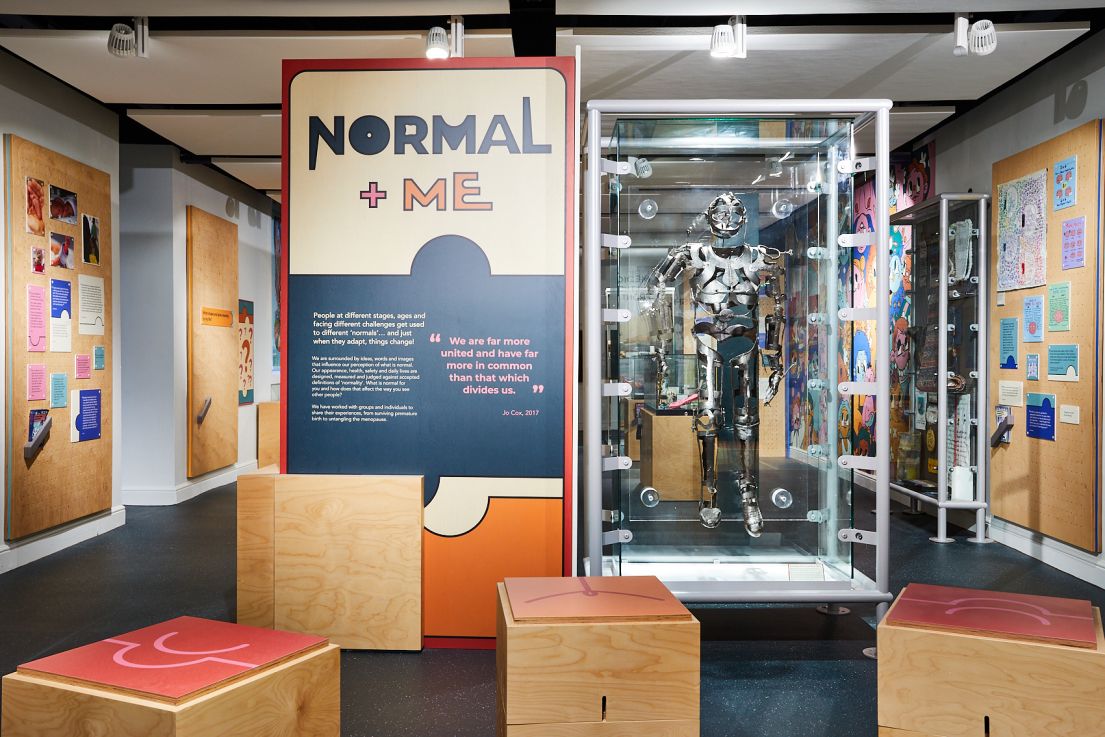 A museum room where there is a metal deconstructed figure in a glass case next to a sign that reads Normal + Me.