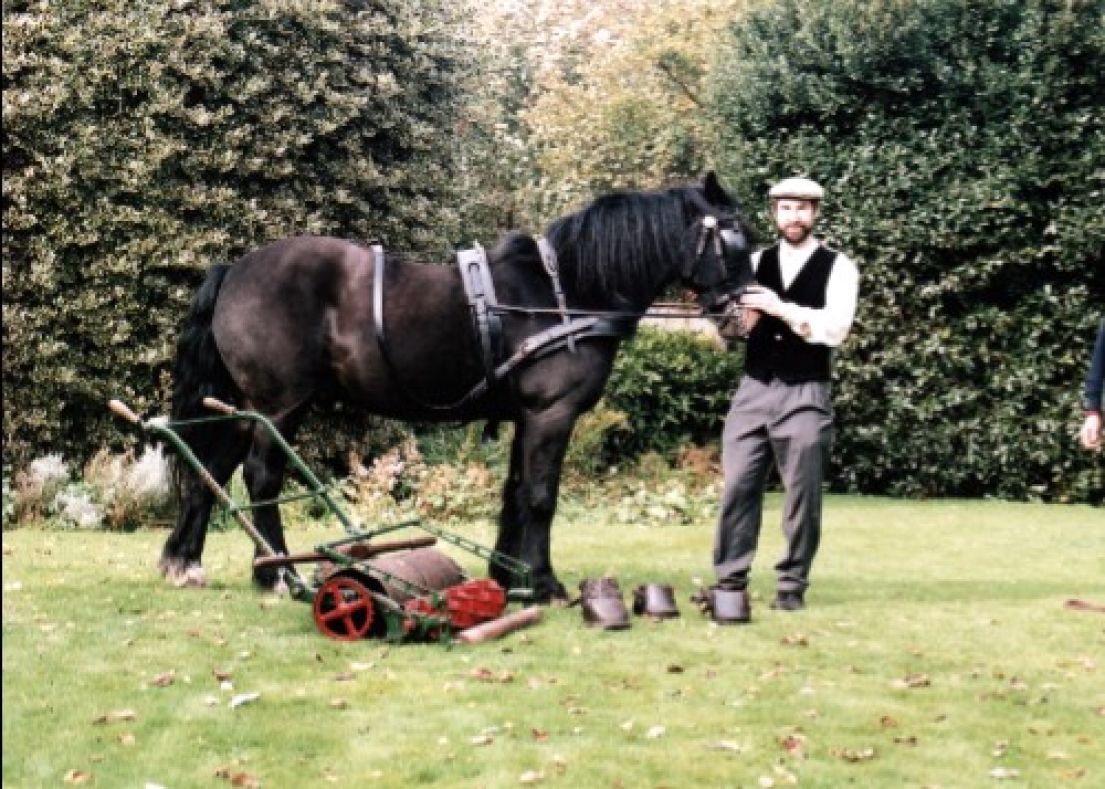 A man holding a dark brown pony by the reigns. By the horses feet is a metal lawnmower and four leather pony boots.