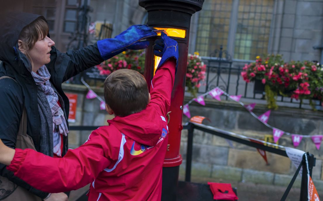 A women and small boy reaching up to post a letter to burn into an incinerator disguised as a letterbox. In the background is HODs bunting.