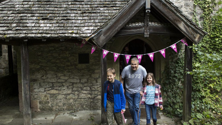 A family with one adult male and two small children excitingly walking out from a church porch adorned in HODs bunting.