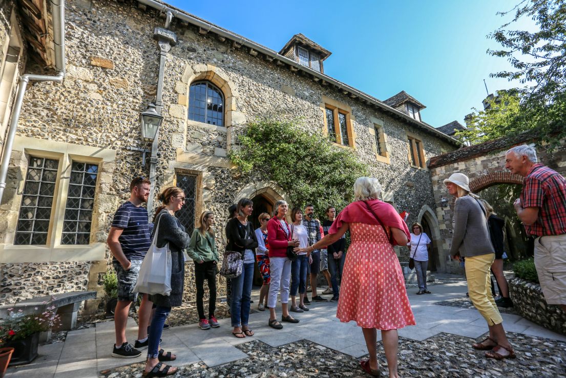 A number of visitors standing around in a oddly formed group in a stone courtyard of a medieval stone building, listening to a person speak.