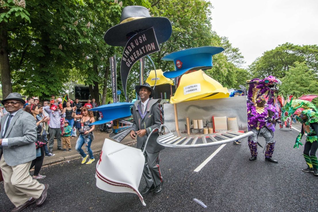 A street parade. A Black man is foregrounded, in a grey suit with large models of a hard hat, fedora, TFL hat and nurses hat attached to him by poles.