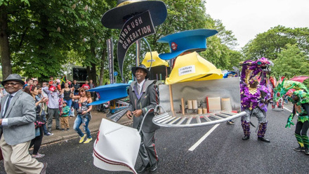 A street parade. A Black man is foregrounded, in a grey suit with large models of a hard hat, fedora, TFL hat and nurses hat attached to him by poles.